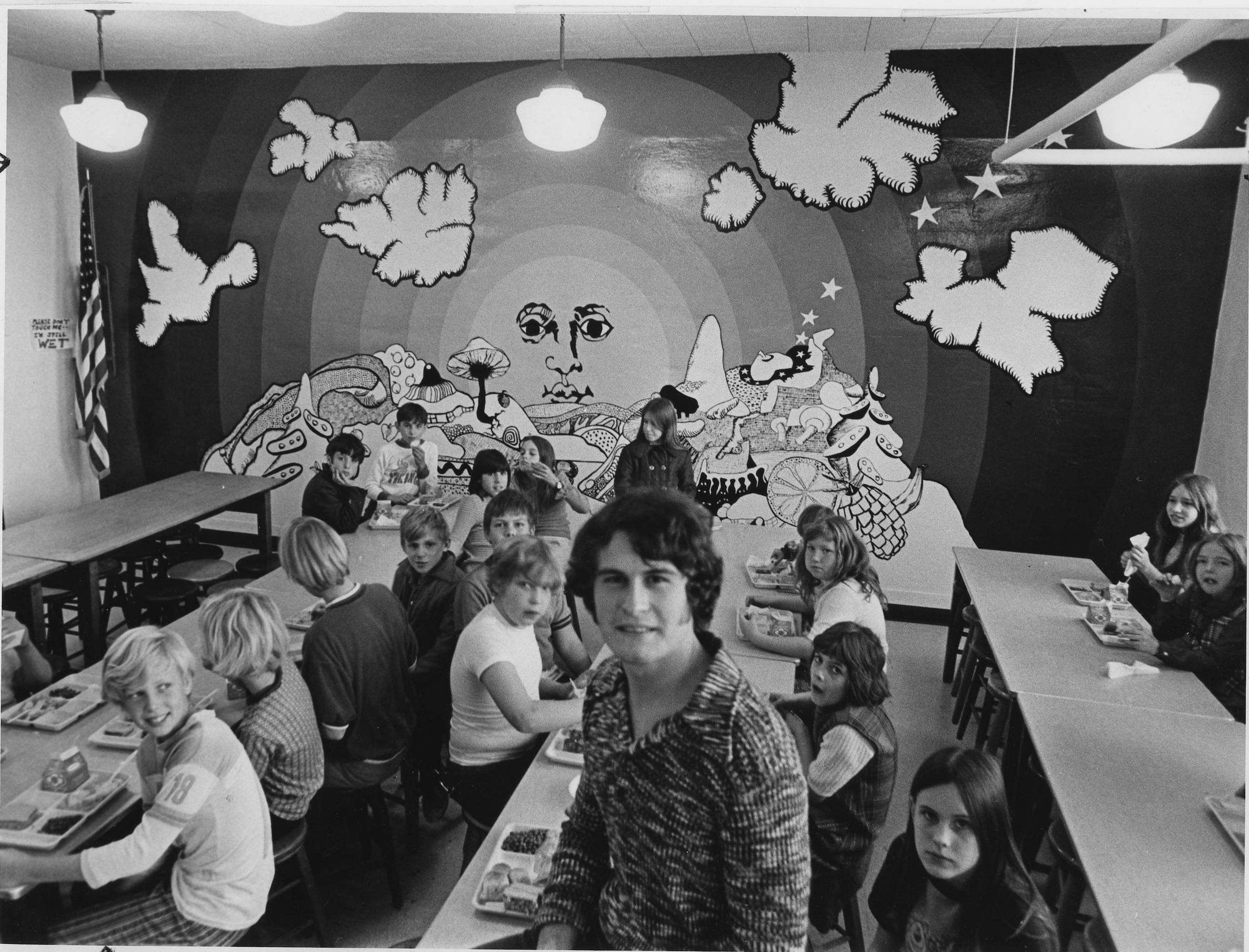 The lunchroom around 1974, with a large mural of a sun with a face on the end wall.
